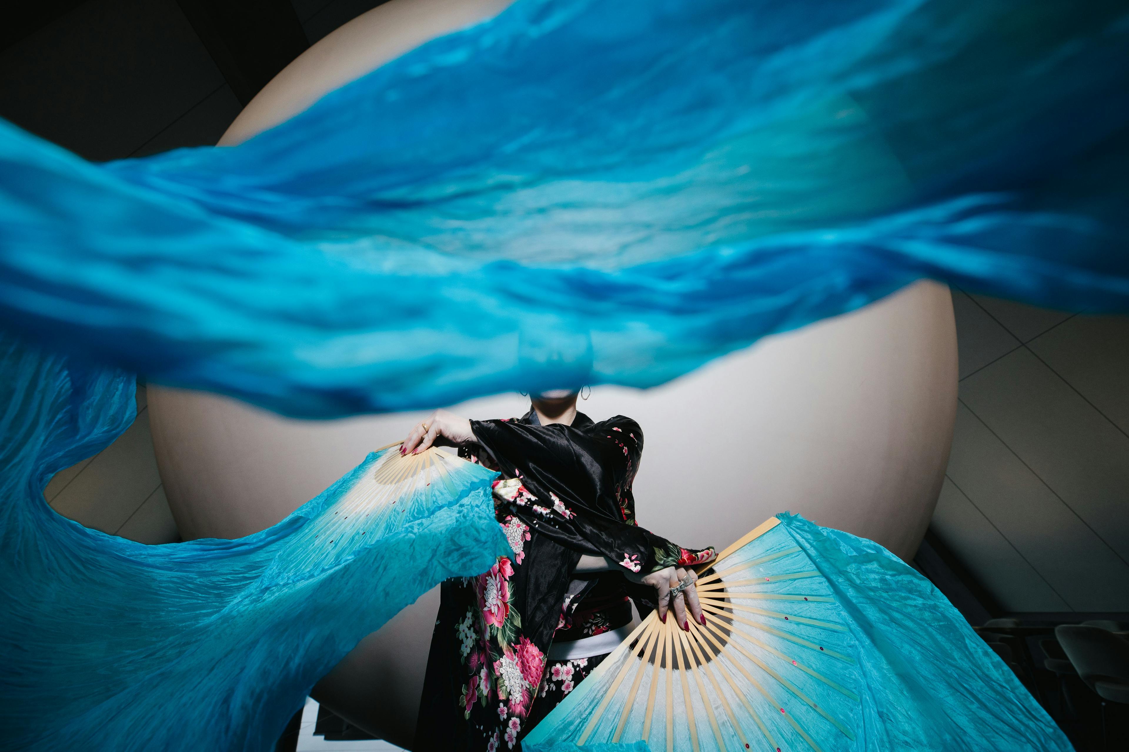 Swatches of blue fabric attached to fans billow in front of the camera held by someone in elegant black silk