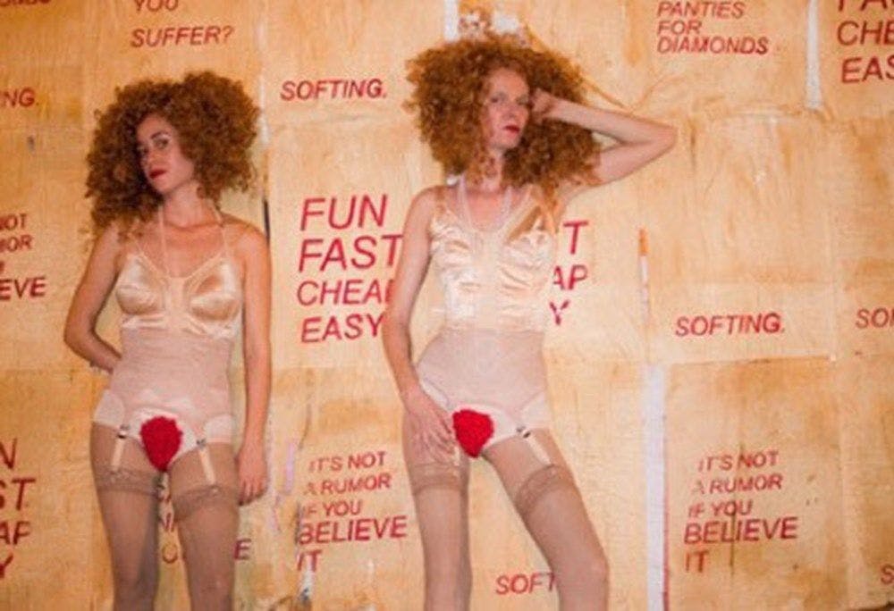 Two women in garters and satin bras with clown red public hair pose in front of a wall with the words "softing" "fun fast cheap east" stenciled in red.