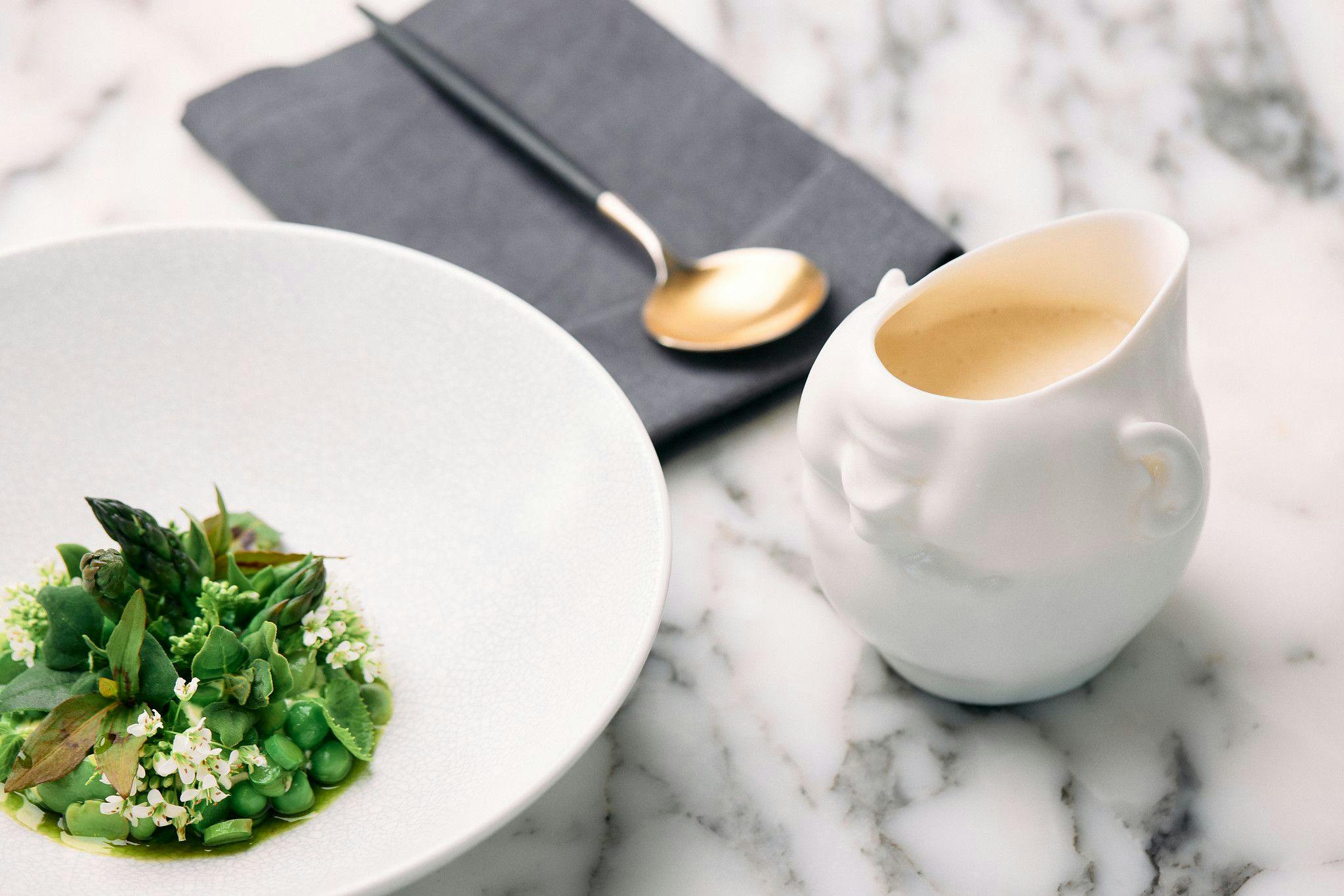 A marble table with a sauce jug of a porcelain upside down baby's head and a plate of vibrant peas, asparagus and delicate white flowers.