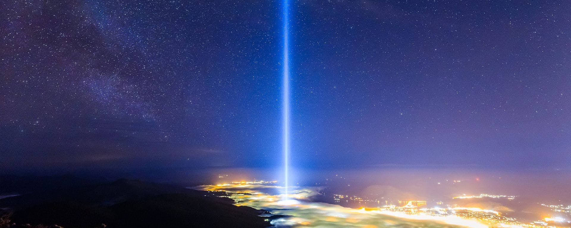 Taken from atop kunanyi or Mount Wellington, a tall beaming tower of blue white light stretches from within a golden sea of the lights of Hobart high up into the night's sky. The sky is dotted all over with starlight.