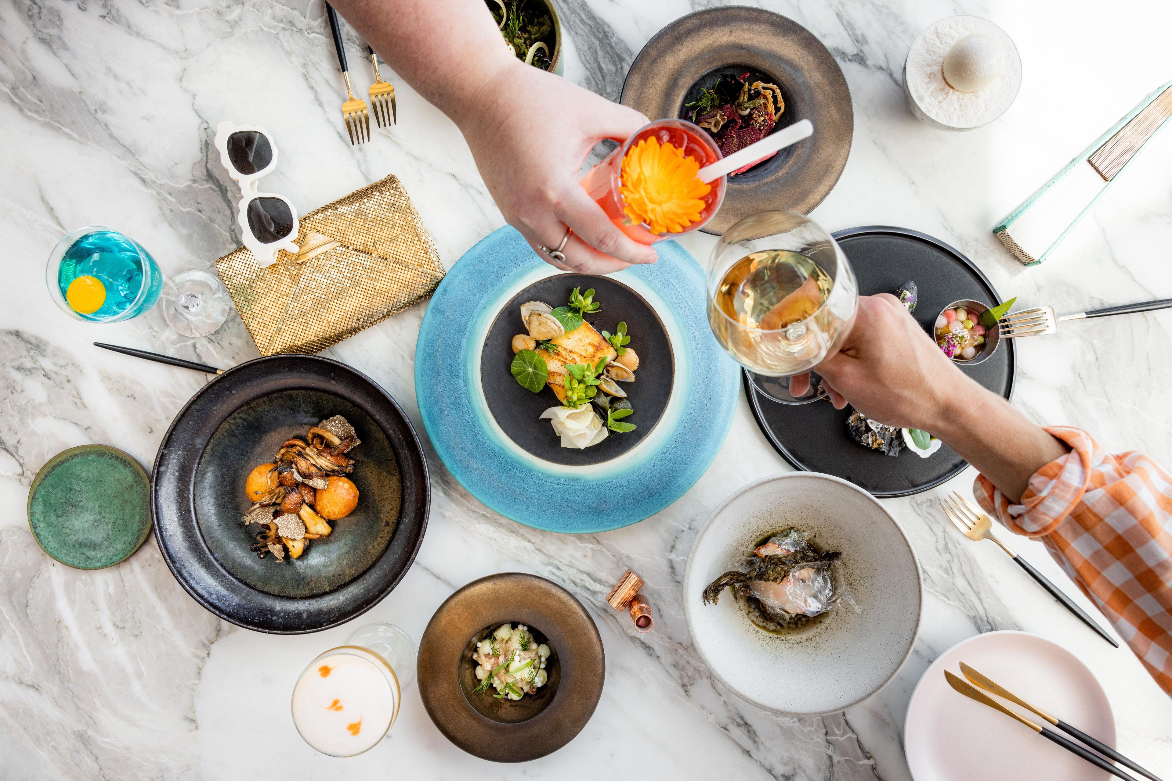 Birds eye view of two people clinking glasses above a marble table laid with delicious looking small bites