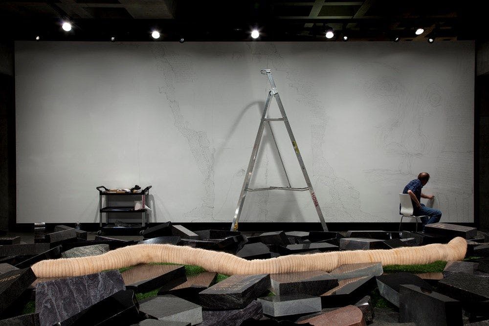 In the foreground lies a large worm on top of black headstones. In the background Jeff Gabel works on a large white canvas.