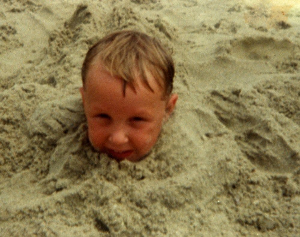 Photo of a small blonde child buried up to their neck in sand at the beach.