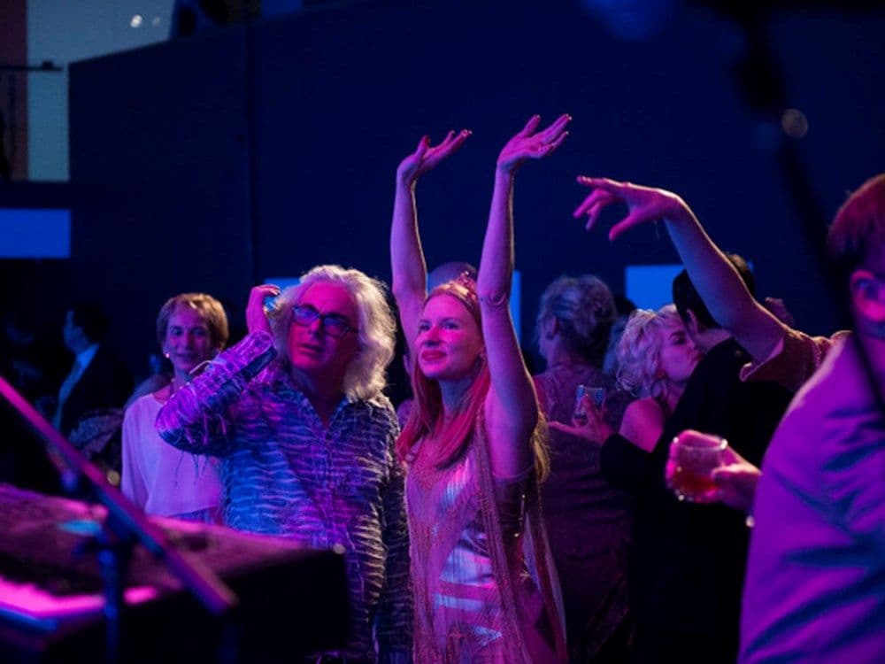 A shot of David and Kirsha dancing to a band performing. Kirsha has both hands raised above her head.